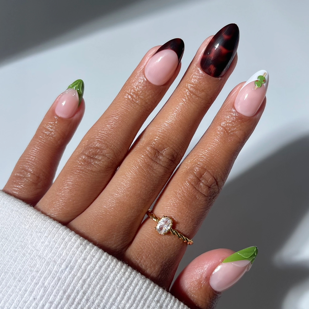 A hand wears glamnetic's Antipasto long almond-shaped press-on nails, showcasing mixed nail art: green leafy tips, pink with white and green accents, and dark marbled nails. A gold ring with a clear stone completes the look.