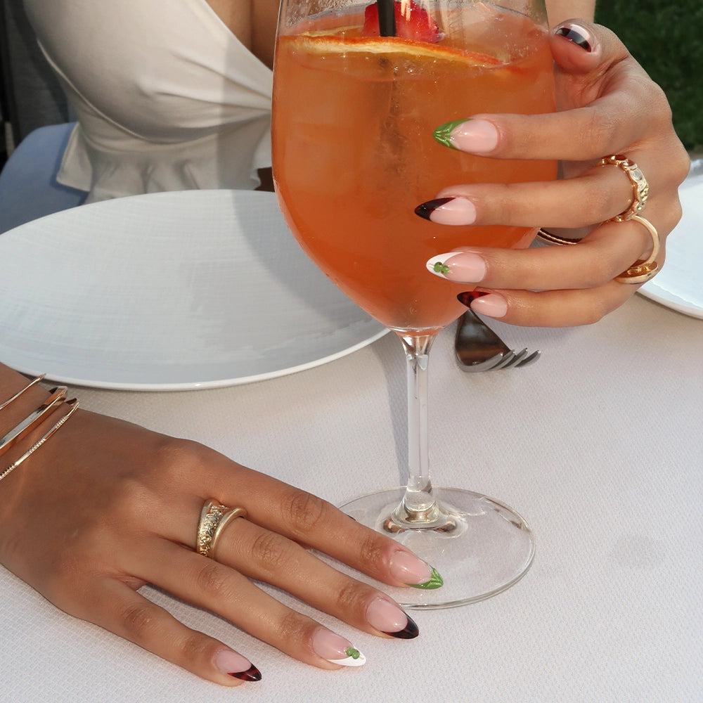 A woman wearing gold rings and bracelets showcases glamnetic Antipasto press-on nails with colorful tips while holding an orange cocktail with a lemon slice at a white-clothed table set with a plate and cutlery. - Product image