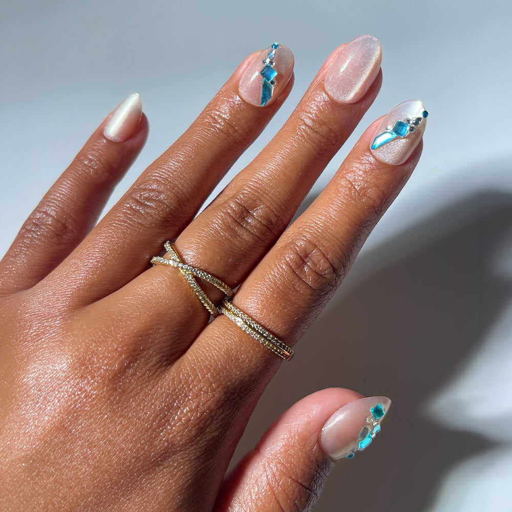 A hand adorned with gold rings showcases manicured short oval nails featuring "Aquamarine" press-on nails by glamnetic, decorated with blue and silver geometric nail art and gemstone-inspired rhinestones, set against a light background.
