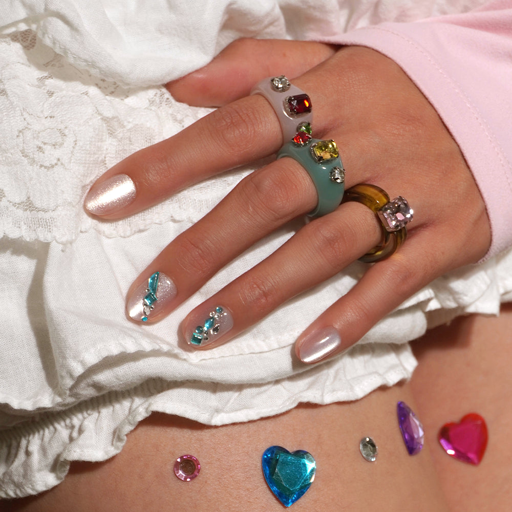 A hand with pastel manicured nails and colorful gemstone rings rests on white fabric. The short oval nails showcase subtle gemstone nail art inspired by the glamnetic Aquamarine press-on nails, while gem-shaped stickers are scattered across the skin below. - Product image