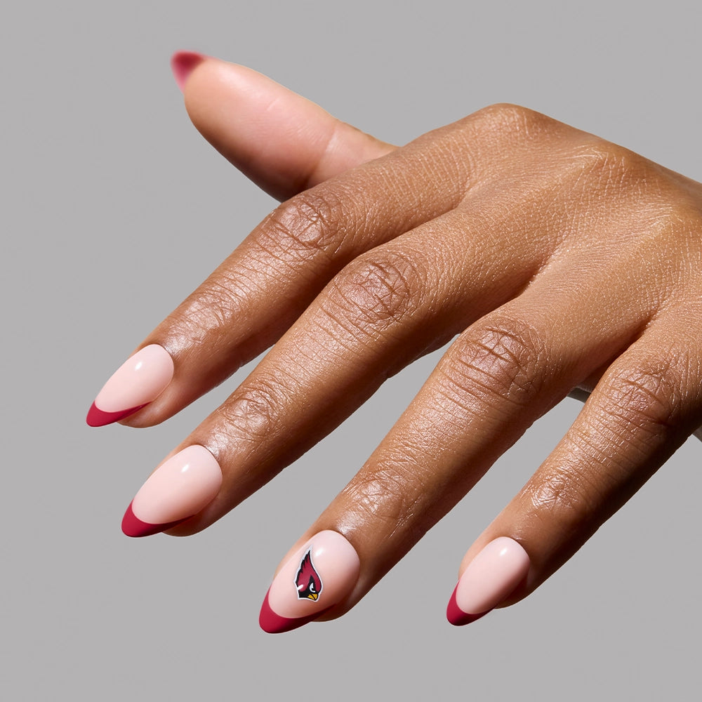 A hand showcases the glamnetic Arizona Cardinals Press-On Nail Set with almond French tips, a light pink base, red tips, and a cardinal bird on the ring fingernail, set against a plain gray background. - Product image