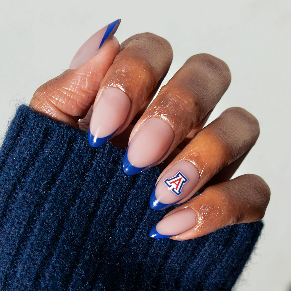 A hand with long nails featuring the glamnetic Collegiate Press-On Nails is painted nude with blue tips. One nail displays a University of Arizona “A” logo, and the person is wearing a dark blue sweater. - Product image