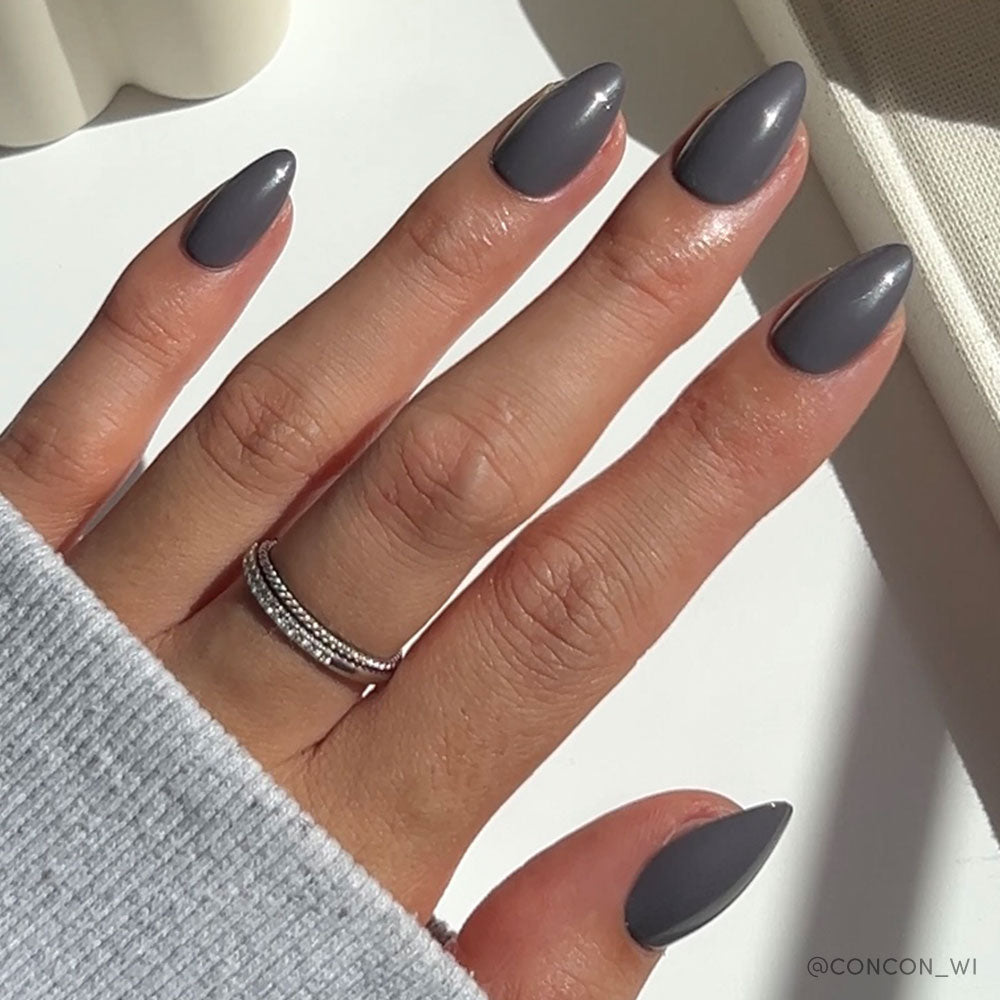A hand with long, almond-shaped Ash Gray nails by glamnetic is polished in glossy grey. The person wears a silver double-band ring on the middle finger and a light grey long-sleeve top, with sunlight casting soft shadows. - Product image