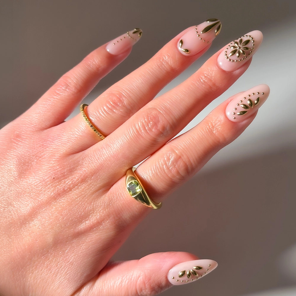 A hand showcases glamnetic's Athenian Gold press-on nails, featuring an almond shape in pale nude with gold accents and delicate patterns, accessorized with a thin gold band and a thicker gold ring set with a small gemstone.