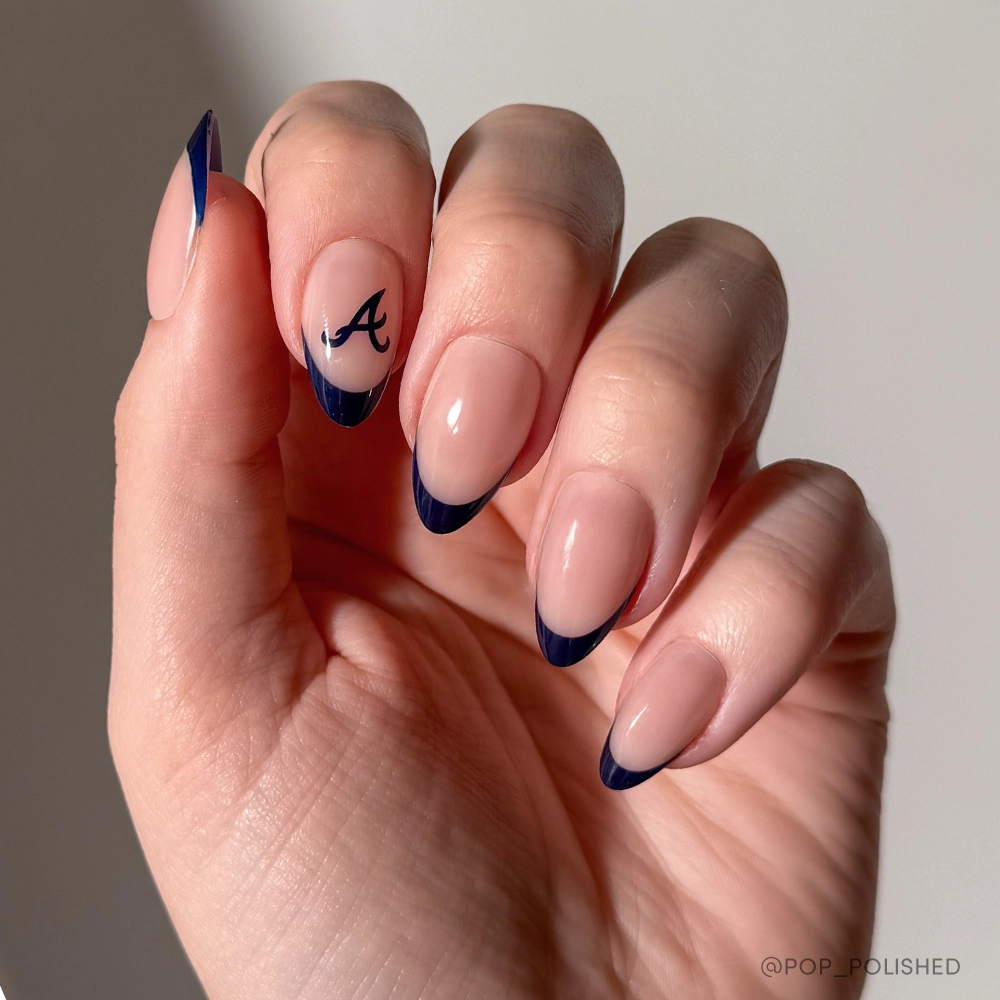 A hand with almond-shaped nails from glamnetics Atlanta Braves Press-On Nail Set: nude base, dark blue French tips, black letter A on the ring finger. Perfect for fans! Background is plain and light-colored. - Product image