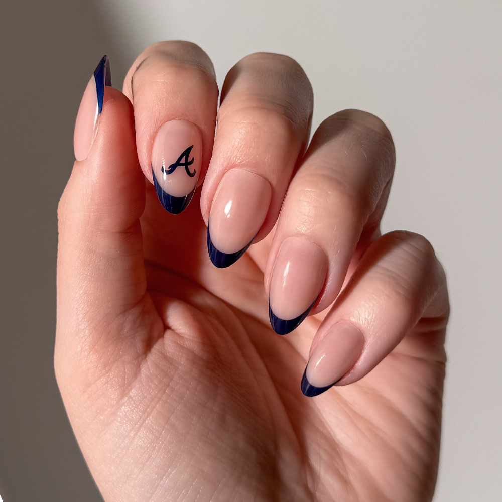 A hand featuring almond-shaped glamnetic MLB Press-On Nails in a nude base shade with dark blue French tips; the ring finger displays a black letter A design near the cuticle. - Product image