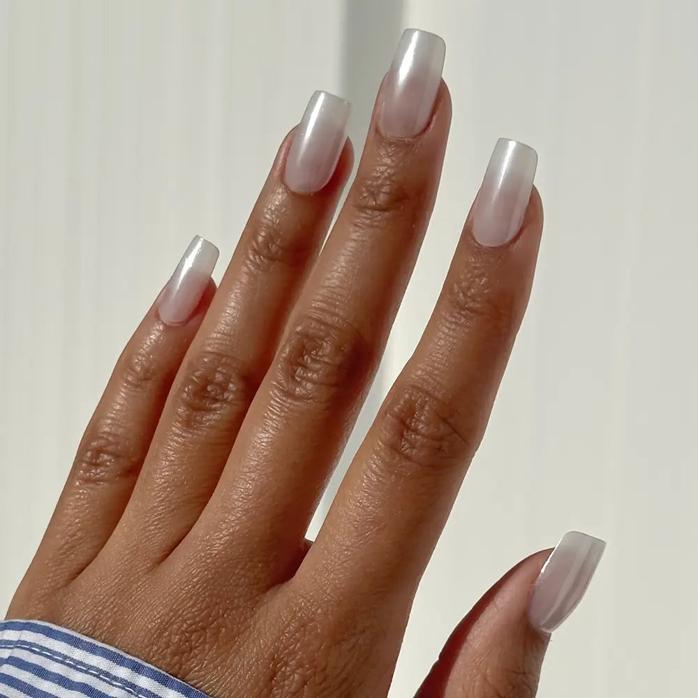 A hand with medium-dark skin displays long, medium square glamnetic “Bailey” press-on nails, painted in a shiny, pearly white polish against a light background.