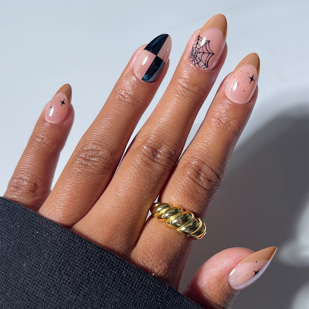 A hand with short almond glamnetic Bat Check press-on nails showcases Halloween-inspired nail art, including spiderwebs, stars, and black geometric designs for a salon-quality manicure. The look is accessorized with a chunky gold ring and a black sleeve, perfectly complementing the Bat Check Press-On Nail Set.