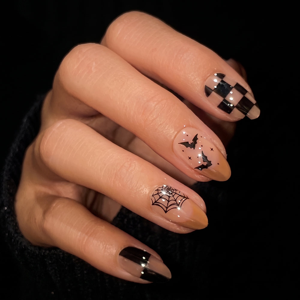 A hand with long nails featuring Halloween-themed nail art: black and nude checkerboards, a spider web, bats, and star designs—just like the Bat Check Press-On Nail Set—against a dark background for a salon-quality mani at home. - Product image