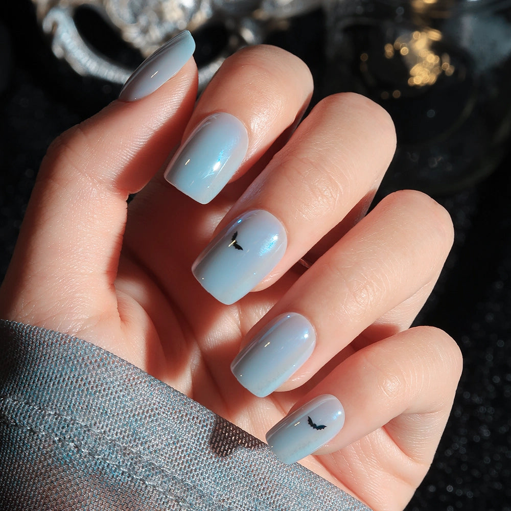 A hand showcases glamnetic’s Batty Bits press-on nails—a set of light blue, glossy short squoval nails, with two accent nails featuring petite black bat-inspired designs. Sunlight highlights their effortless charm, while a blue sleeve partially covers the wrist for an extra touch of chic.