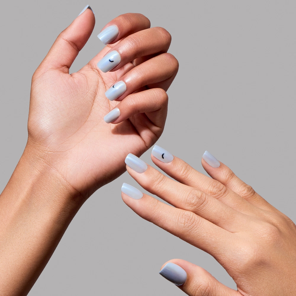 A person with medium skin tone shows off short squoval nails manicured in light blue using glamnetic’s Batty Bits. Their left hand features a small black accent mark on the middle fingernail, resembling subtle bat nail art. The background is plain gray. - Product image