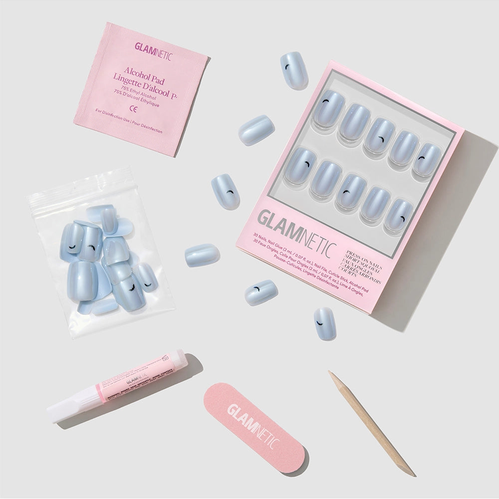 A glamnetic Batty Bits nail kit is displayed on a gray background, featuring light blue Short Squoval Press-On Nails adorned with small eyelash designs. The stylish nails come packaged with essential accessories, including nail glue, an alcohol pad, a pink nail file, and a wooden cuticle stick. - Product image