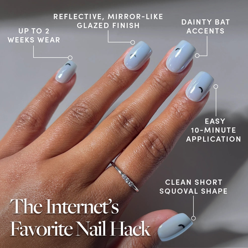 A hand with light blue, glazed short squoval press-on nails from glamnetic’s Batty Bits set, featuring small bat nail art accents. Text highlights easy 10-minute application, reflective finish, and up to 2 weeks of wear. The hand is accessorized with a silver ring. - Product image