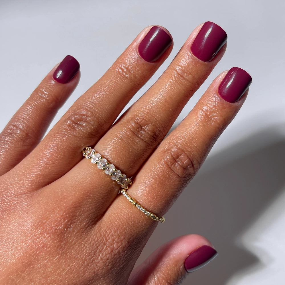 A hand with super short, glossy maroon Berry Baby nails by glamnetic wears two gold rings—one featuring clear gemstones and another plain band—against a light background.