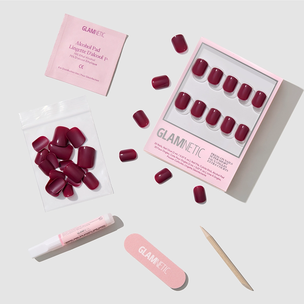 A flat lay of the glamnetic Berry Baby press-on nail kit featuring maroon nails, glue, alcohol pad, nail file, cuticle stick, and extra nails arranged neatly on a light gray background.