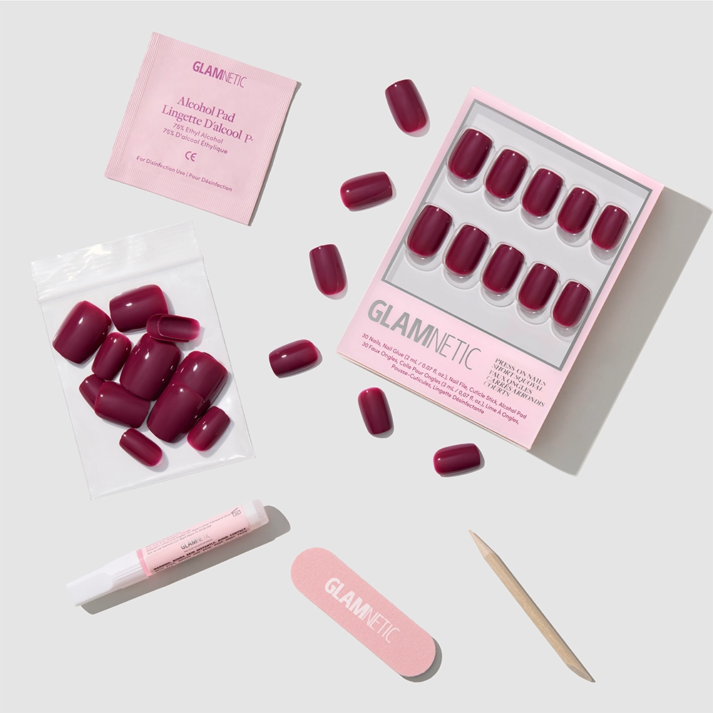 A flat lay of glamnetic Berry Crush, a boxed set featuring short squoval press-on nails in a deep berry hue with loose nails, nail glue, an alcohol pad, a pink nail file, and a wooden cuticle stick.