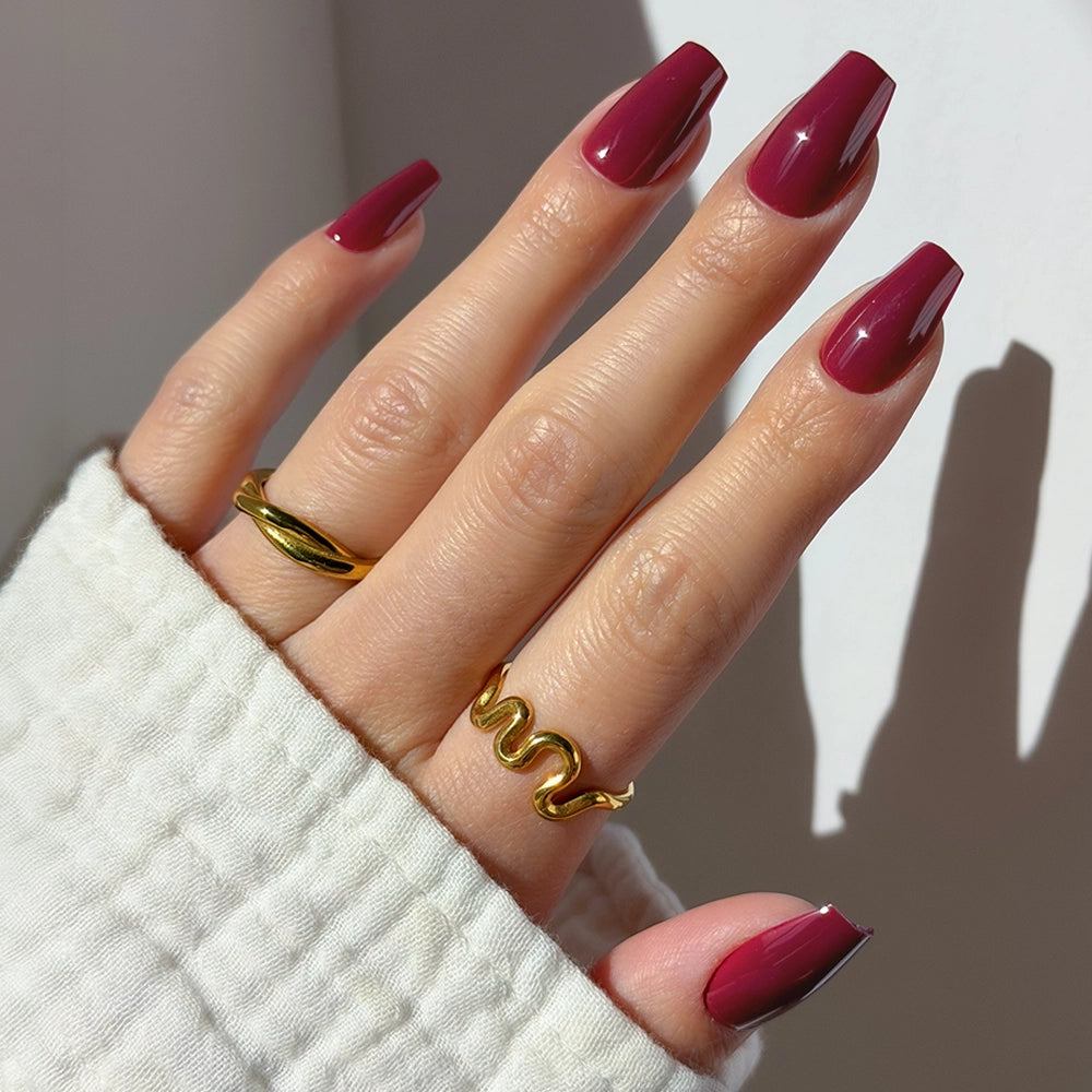A hand with glossy maroon Berry Fierce press-on nails from glamnetic in a coffin shape wears two gold rings—one twisted, one wavy—against a white textured sleeve and a neutral background.