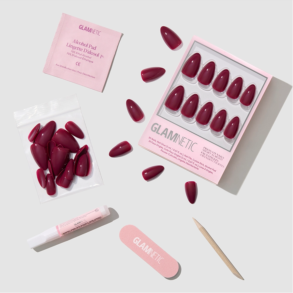 A set of glamnetic Berry Kiss maroon press-on nails in a short almond shape, shown with an alcohol pad, nail glue, nail file, wooden cuticle stick, and extra nails, neatly arranged on a light gray background.