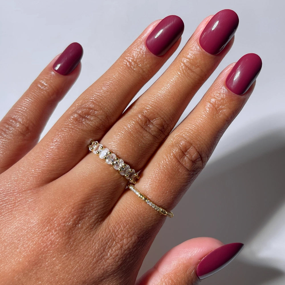 A hand with glamnetic’s Berry Maroon oval press-on nails and two gold rings—one with clear gemstones, one with small diamonds—rests against a plain background, showcasing a chic autumn-ready look.