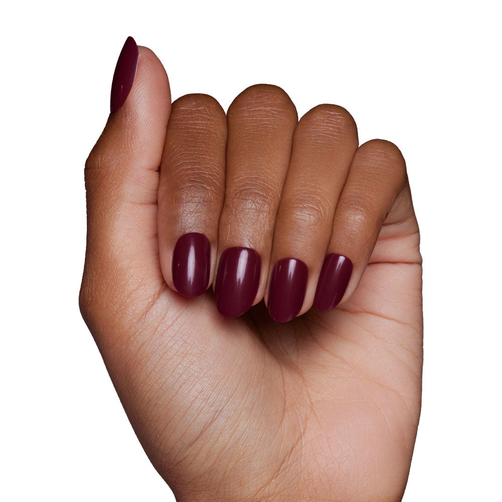 A hand with medium brown skin tone displays oval-shaped glamnetic Berry Maroon press-on nails, the glossy maroon color standing out as fingers curl inward against a crisp white background. - Product image