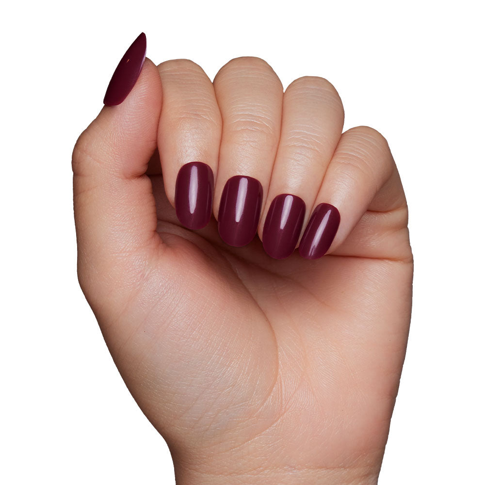 A hand with glamnetics Berry Maroon press-on nails, featuring medium-length oval tips in a glossy dark burgundy shade, is displayed against a plain white background. - Product image
