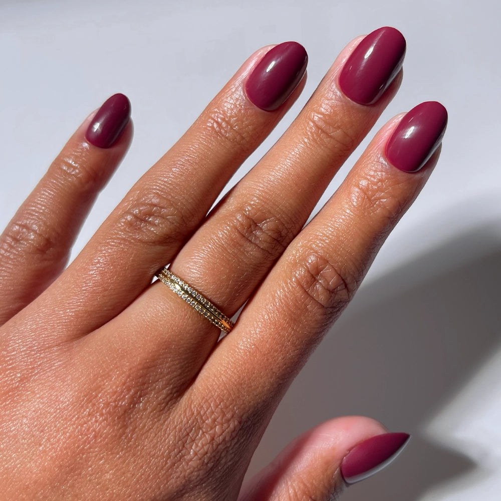 A hand with medium skin tone displays neatly manicured short oval nails painted in glamnetic's Berry Mood, a glossy maroon shade. A thin gold ring with small stones adorns the ring finger. The background is white.