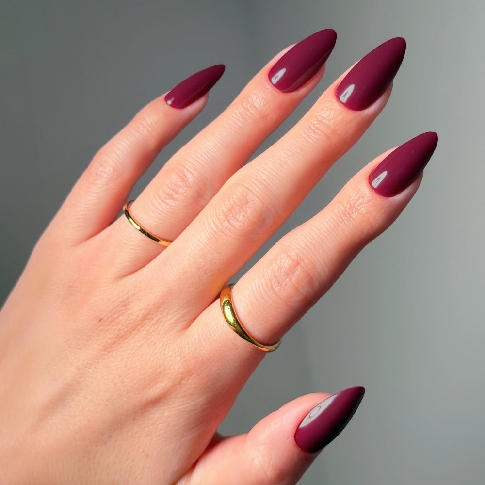 A hand with long, almond-shaped glamnetic Berry Obsession press-on nails in glossy maroon rests against a soft, neutral background. Two simple gold rings on the middle and ring fingers highlight the elegant manicure.