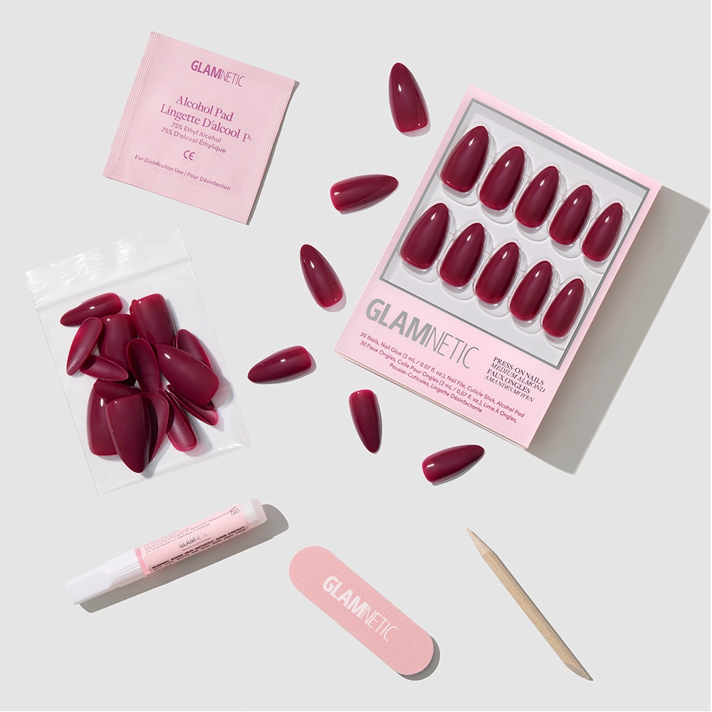 The Berry Obsession set by glamnetic features maroon almond-shaped press-on nails, a pink nail file, wooden cuticle stick, alcohol pad, adhesive tube, and extra nails, all elegantly displayed on a light gray background.