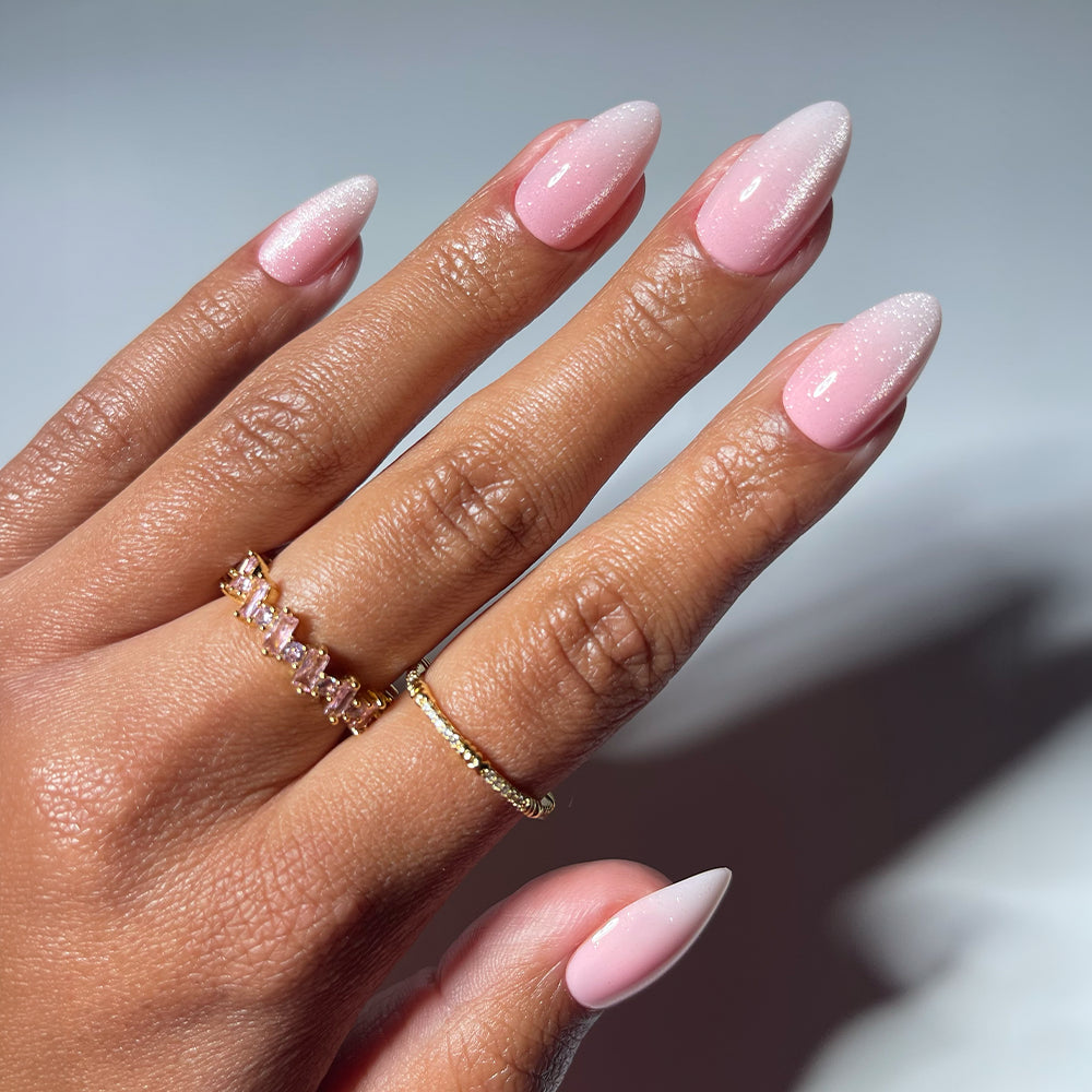 A hand displays glamnetic's BIG BUNNY ENERGY almond-shaped press-on nails in a glossy baby pink ombre, complemented by two gold rings—one with rectangular stones and one plain—in a softly lit setting that highlights their elegance.