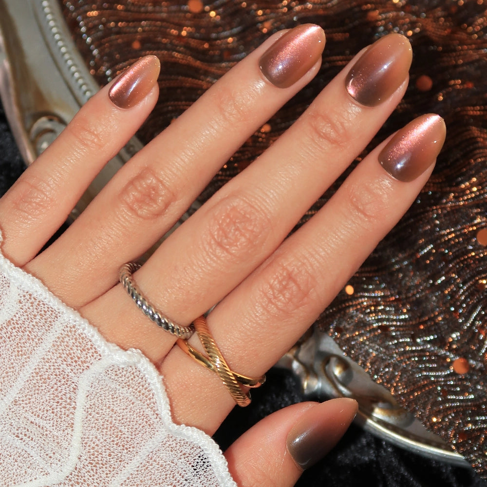 A hand decorated with glamnetic’s Bitter Orange short oval press-on nails in a lustrous, brownish metallic shade, paired with two gold rings and a white lace sleeve. The background showcases a sparkling sequined fabric for added glamour.