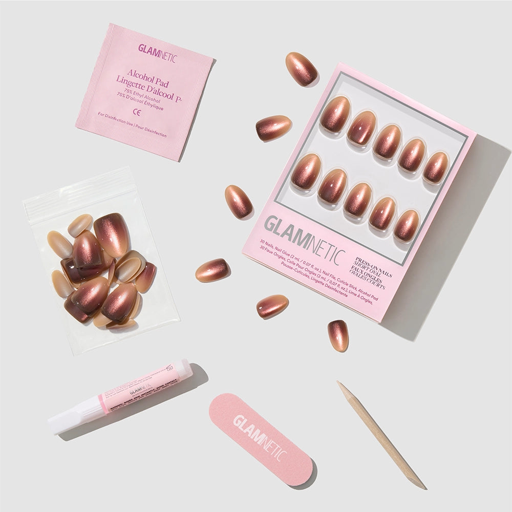 The “Bitter Orange” press-on nails set by glamnetic showcases short oval nails in a luxurious metallic rose gold finish and includes adhesive, an alcohol pad, nail file, wooden cuticle stick, and extra nails—all neatly arranged on a white background. - Product image