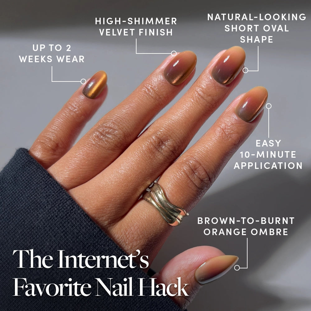 A hand with short, oval nails displays a high-shimmer, brown-to-burnt orange ombre manicure created using the glamnetic Bitter Orange Press-On Nails. These nails provide long wear, easy application, and a velvet finish. The look is accessorized with two gold rings. - Product image