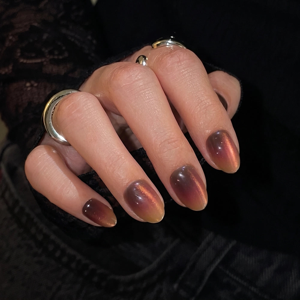 A hand with almond-shaped nails adorned in a shimmery, gradient dark brown and copper polish, reminiscent of the glamnetic Bitter Orange Press-On Nail Set. The person accessorizes with silver rings and a black lace sleeve. - Product image