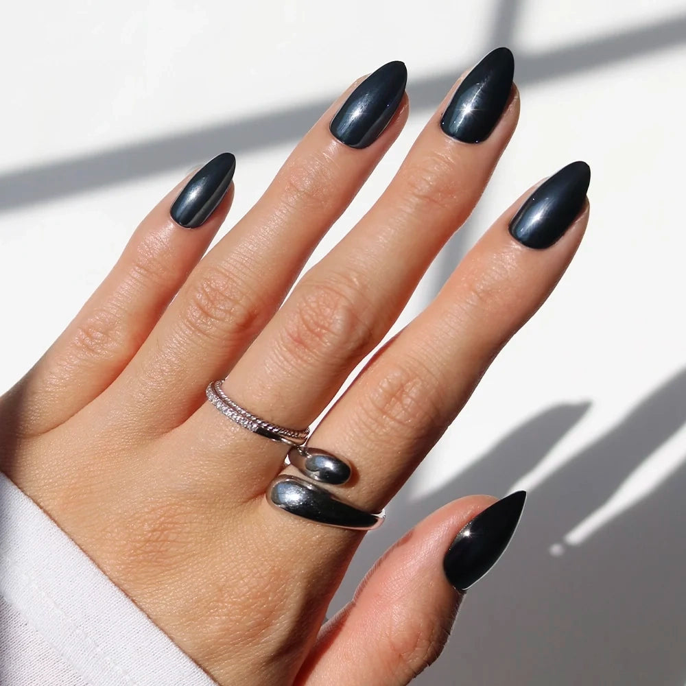 A hand featuring long, almond-shaped Black Metal press-on nails by glamnetic, with two silver rings adorning the middle and ring fingers, is displayed against a white background. These vegan and cruelty-free nails deliver a striking, glossy black chrome finish for an ethical yet glamorous beauty statement.