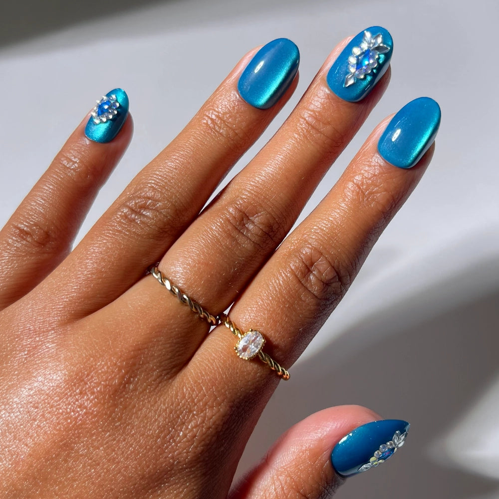 A hand showcases "Blue Topaz" press-on nails by glamnetic in a metallic blue, short oval shape, with some nails adorned with delicate white and silver floral accents. The hand is accessorized with two gold rings—one set with a gemstone—against a neutral background.