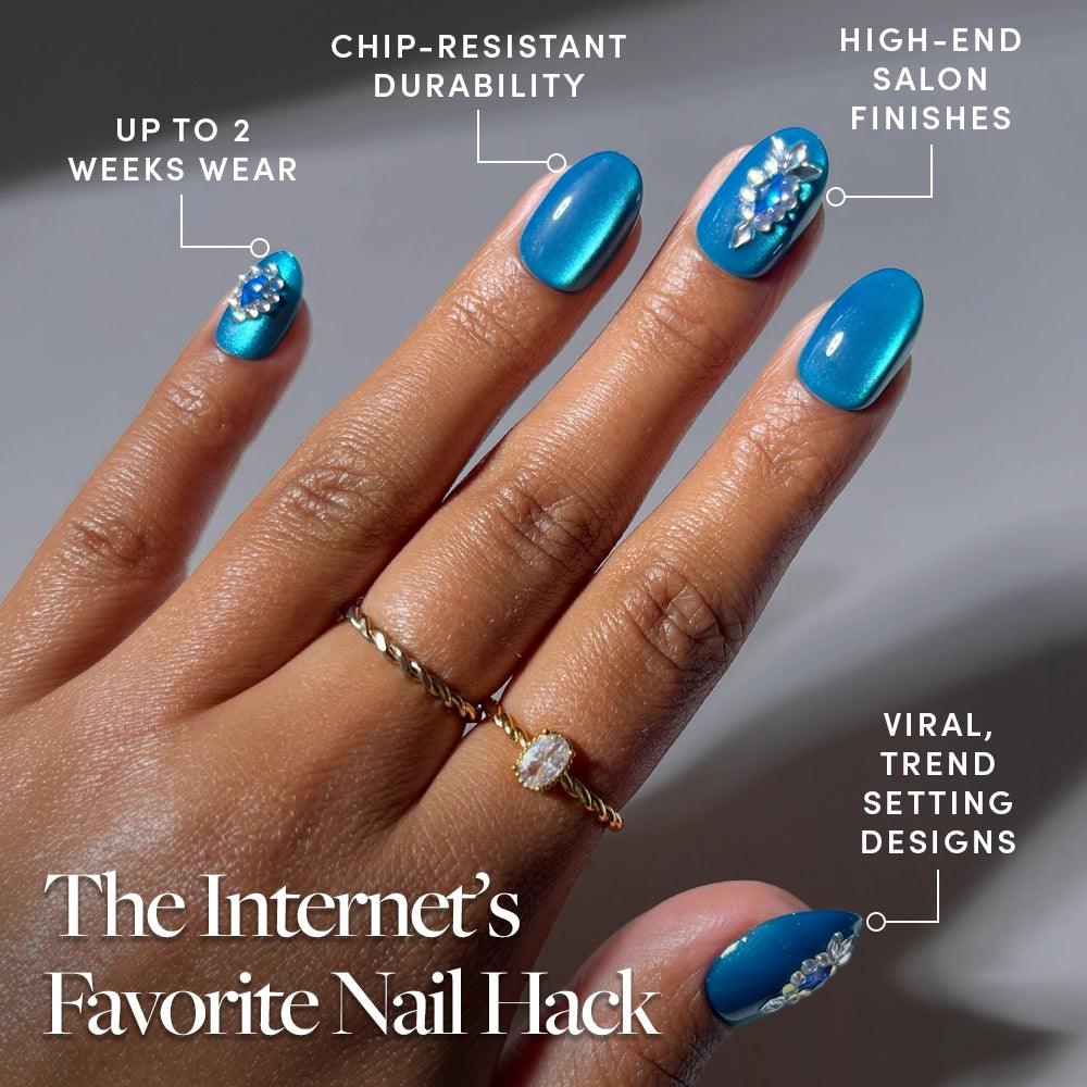 A hand showcases the Blue Topaz press-on nails by glamnetic—short oval nails featuring blue floral designs and gemstone-inspired shine. Text highlights their up to 2 weeks wear, chip-resistant durability, high-end salon finishes, and viral, trendy style. Two fingers wear rings to accentuate the glamnetic Blue Topaz nails’ brilliance. - Product image