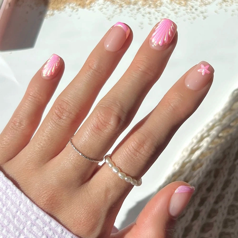 A hand with short squoval nails featuring glamnetic’s “Blush Dunes” press-ons—pink seashell and starfish-themed nail art with white and pink details on a nude base. The look is complete with a pearl ring and a thin silver band.