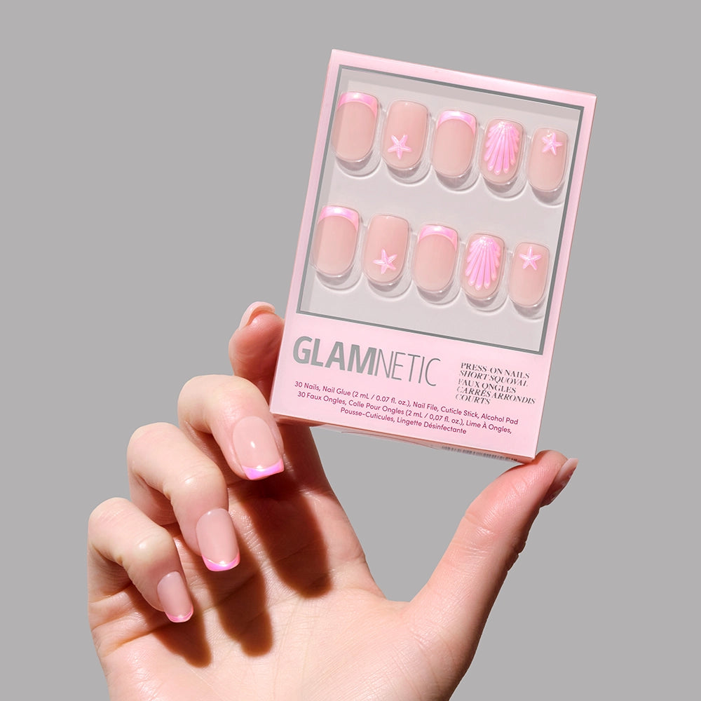 A hand with glossy pink French tips holds a box of glamnetic’s Blush Dunes Press-On Nail Set, featuring soft pink chrome tips and seashell accents, against a plain gray background.