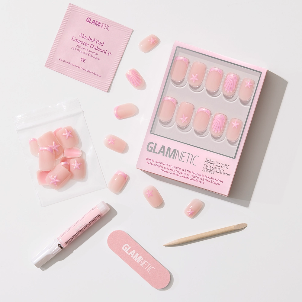 The Blush Dunes press-on nail set by glamnetic features soft pink chrome tips with white star and shell designs, arranged with nail tools on a light background. Packaging displays the glamnetic logo—perfect for tropical getaways. - Product image