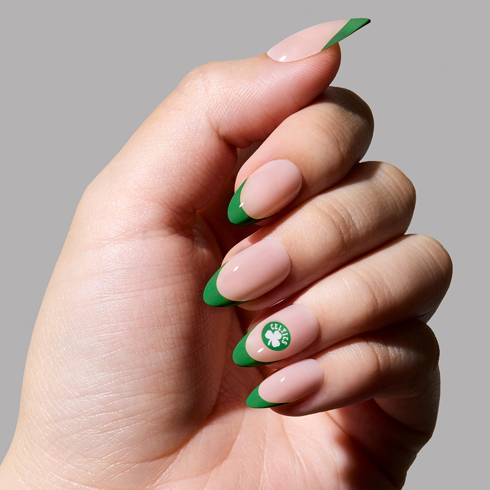A hand displays glamnetic's Boston Celtics Press-On Nail Set: almond-shaped French tips with a nude base, green tips, and a white four-leaf clover on the ring finger. The plain gray background highlights this Fanatics collaboration.