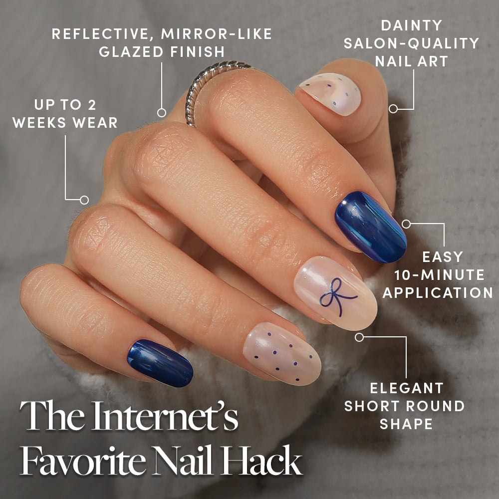 Bows & Bits - A hand wearing Bows & Bits press-on nails with short, round, dark blue and translucent designs featuring dots and a bow, with text callouts highlighting a reflective finish, two-week wear, and salon-quality ease of application.