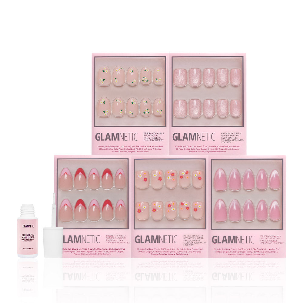 Five boxes of glamnetic Budding Romance pink-themed Glossy Nail Set press-ons, featuring short almond nails, are arranged in two rows. A small open bottle of nail glue is placed beside the boxes on a white background. - Product image