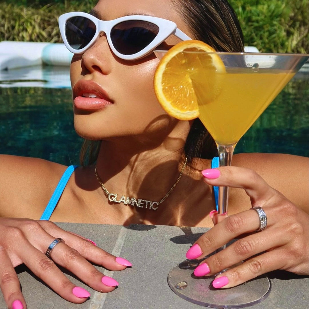 By a pool, a woman in sunglasses and a blue swimsuit holds a martini with an orange slice, flaunting glamnetic Hot Pink nails in a short oval shape, accessorized with rings and a glamnetic necklace amid lush greenery. - Product image