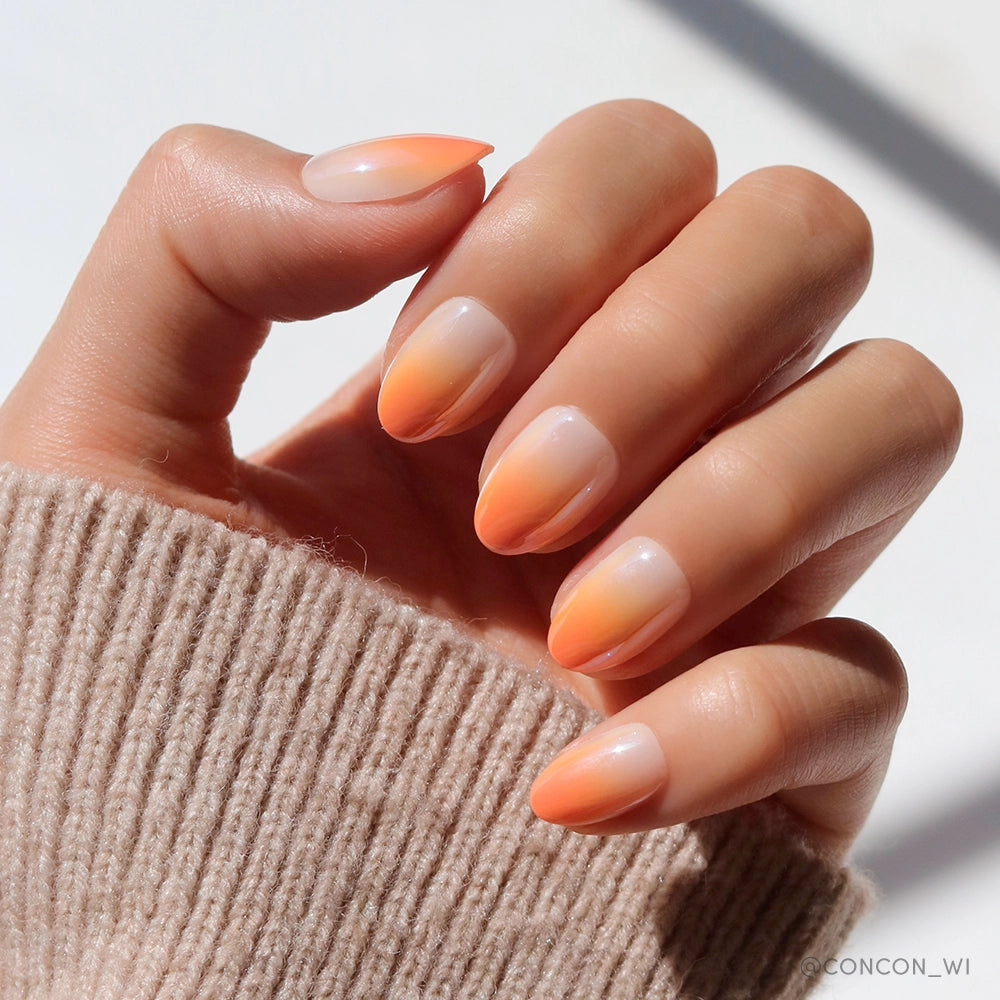 A hand with almond-shaped Candy Corn nails by glamnetic, featuring an orange ombré fade, rests on a beige ribbed sweater—a chic Halloween-ready manicure.