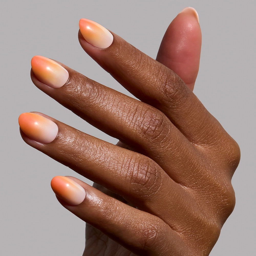 A hand with medium brown skin displays glamnetic’s Candy Corn Nail Set, featuring smooth orange and white ombré, almond-shaped nails that evoke festive Halloween style, against a plain light gray background. - Product image