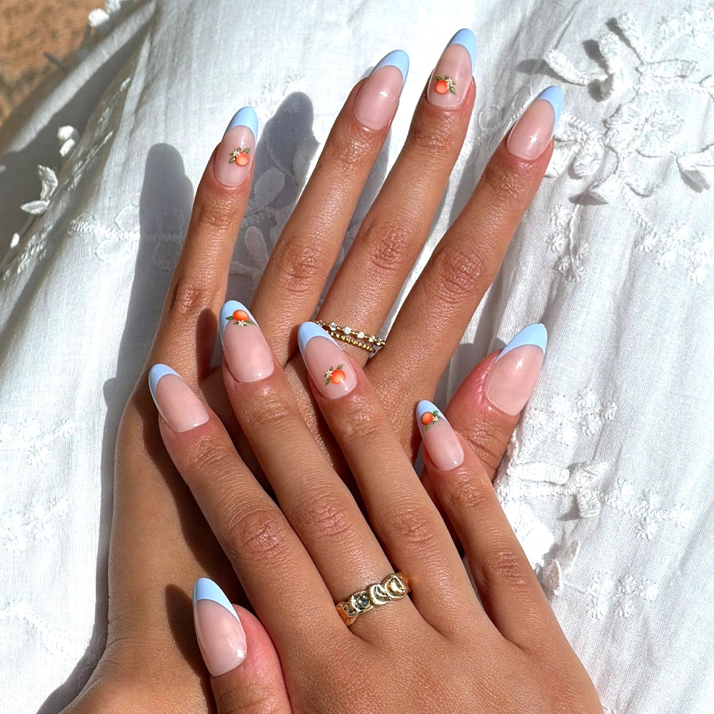 A close-up of hands with glamnetic’s Capri Coast press-on nails, featuring light blue French tips and tiny orange fruit art, rests on white embroidered fabric. Gold rings accent the salon-quality manicure. - Product image