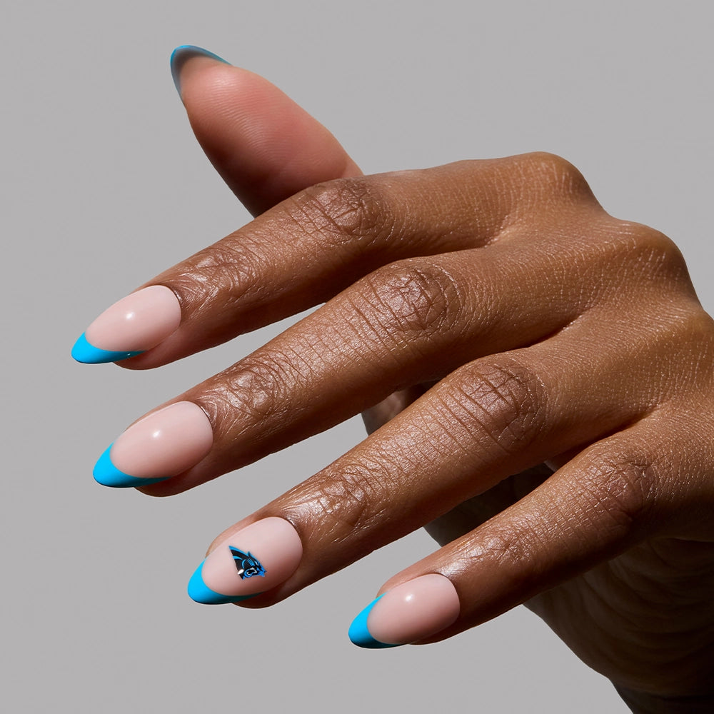 A hand showcases the glamnetic Carolina Panthers Press-On Nail Set, featuring almond-shaped nails with light pink polish, bold blue French tips, and a small blue accent. The background is plain gray. - Product image