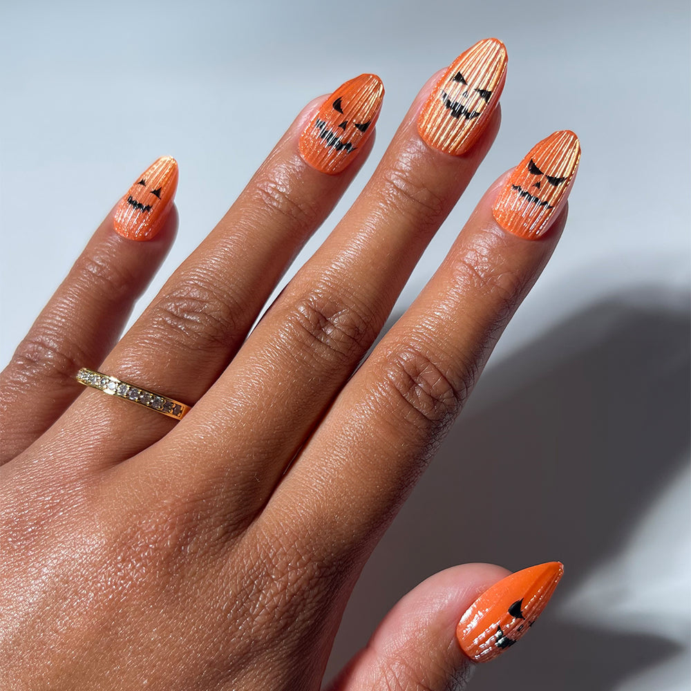 A hand features long, pointed nails adorned with glamnetic’s “Carving Contest” Halloween-themed pumpkin nail art design, showcasing bold orange polish with vertical white stripes and black jack-o-lantern faces.
