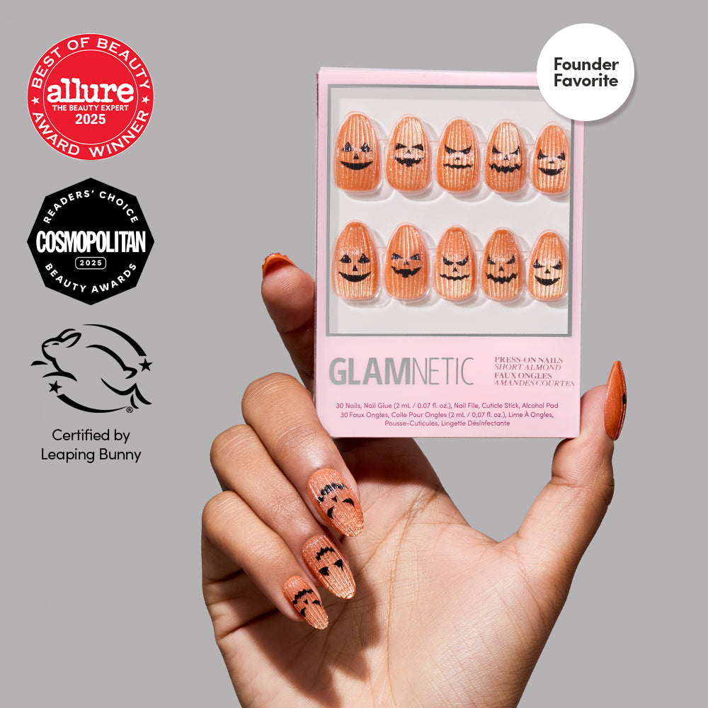 A hand with orange velvet finish Carving Contest press-on nails by glamnetic, featuring jack-o-lantern faces, holds a box of the same pumpkin nail art set. The image also showcases beauty award badges and a cruelty-free certification. - Product image