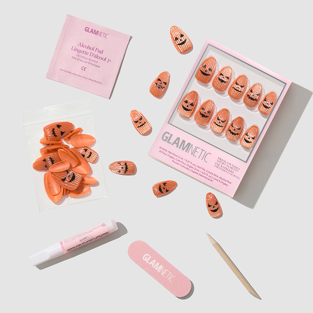 The Carving Contest Halloween press-on nails kit by glamnetic includes orange nails with black jack-o-lantern faces, a nail file, wooden cuticle stick, tube of glue, alcohol pad, and extra nails—all arranged neatly on a light gray background. - Product image